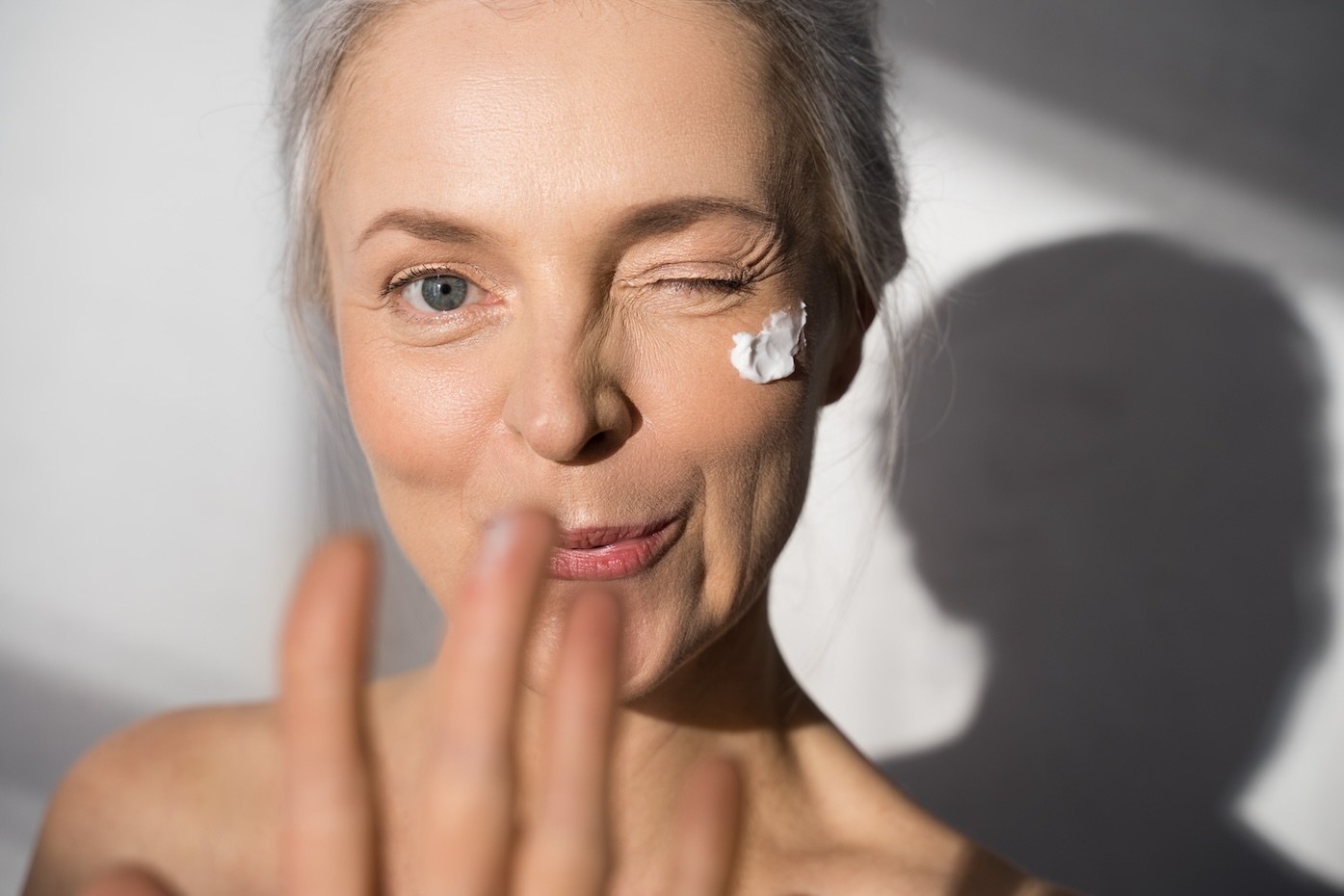 Portrait view of the friendly mature 60-year old woman with protective skin care cream and moisturizer lotion on her happy smiling face posing at the studio and showing her hand to the camera